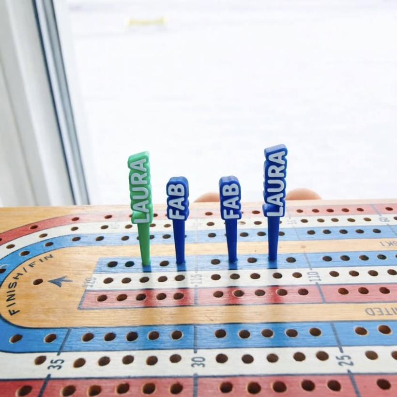 Personalized cribbage pegs with the names LAURA and FAB, in green and dark blue, in a wooden cribbage board